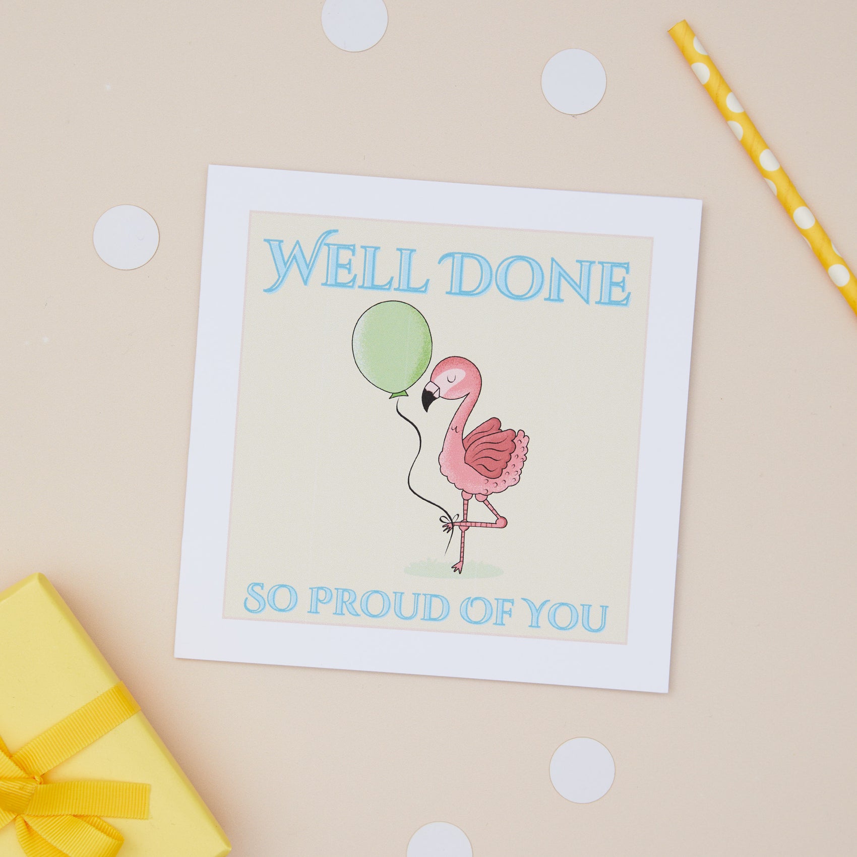 Kids Well Done Card featuring a Pink Flaming holding a balloon. The Card Features the text "well done - so proud of you". The Pink Flamingo sits in the middle of the card, and is holding a green balloon. The Card sits on a beige background, with a present and pencil in the background 