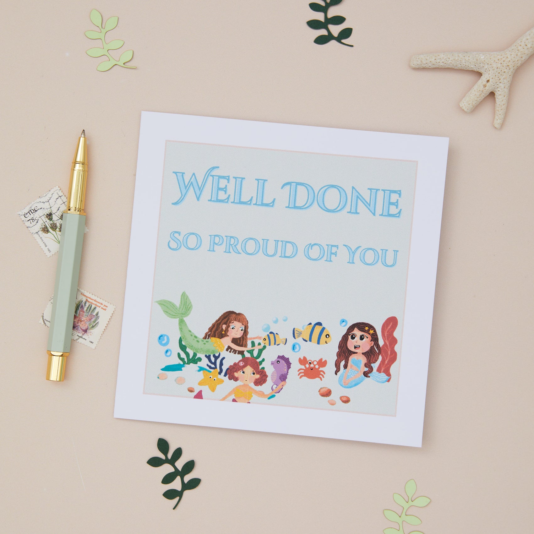A Kids 'Well Done' greeting card with a mermaid theme, featuring illustrations of mermaids, fish, and aquatic creatures. The card is accompanied by a stationery items and a gold pen, displayed on a neutral background with decorative elements like leaves and a starfish.