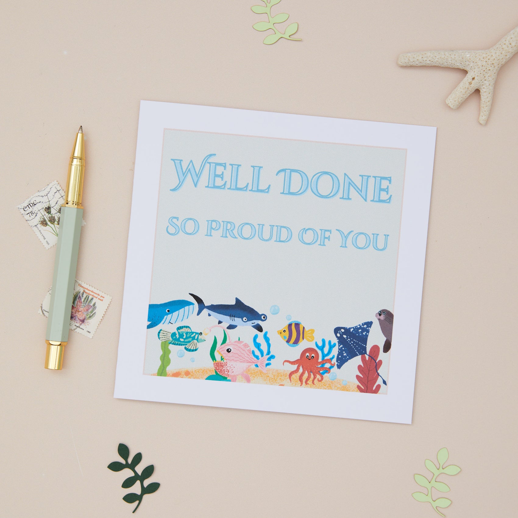 A kids well done greeting card with a 'Well Done' message and a playful sea life design, including a crab, fish, and whale, accompanied by a gold pen on a table with some decorative leaves.