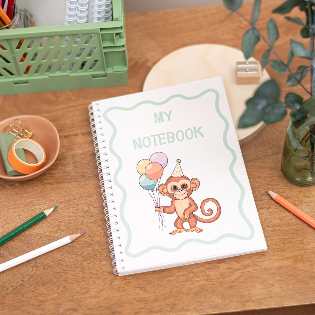 Kids Wirebound Party Monkey A5 Notebook. A handmade A5 notebook with a cover featuring an illustration of a monkey holding balloons, alongside the text 'MY NOTEBOOK'. The notebook is displayed on a wooden table alongside pencils and other items.