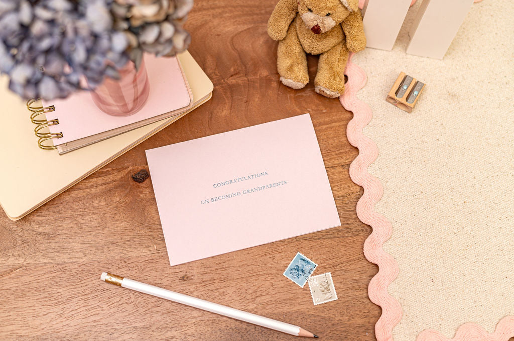 Congratulations on becoming grandparents in silver font on a  pink letterpress card. The card is set on a desk with stationery items around.