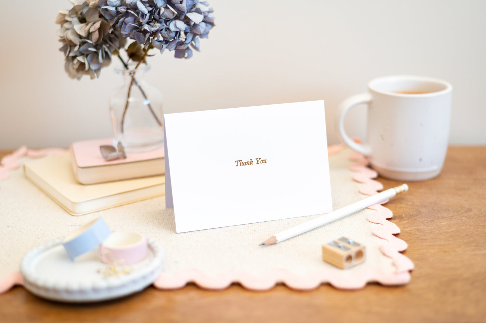 A folded 'Thank You' letterpress card on a wooden table with a cup of tea, a flower vase, and other stationery items in the background.