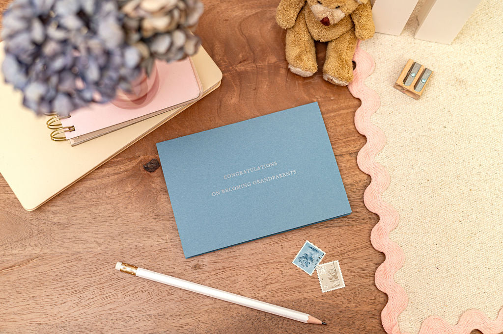 Letterpress Congratulations on becoming grandparents on blue card. The card is set on a desk with stationery items around it.