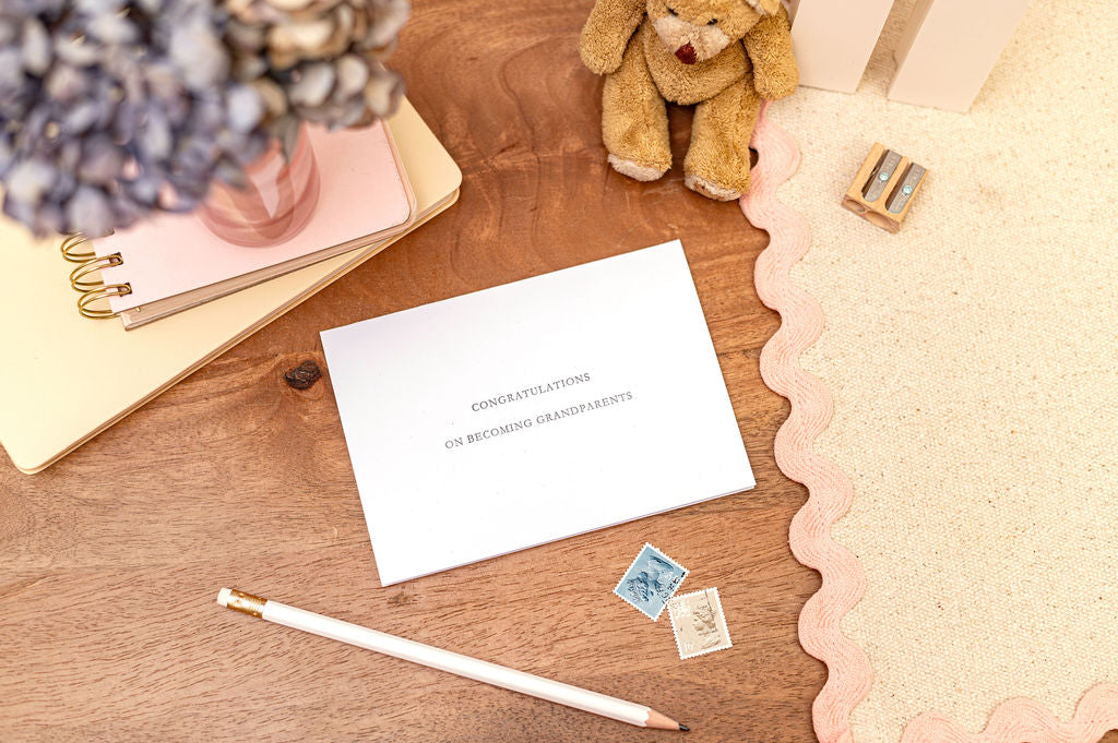 Letterpress congratulations on becoming grandparents card with the words in silver. Card is set against a desk with stationery items