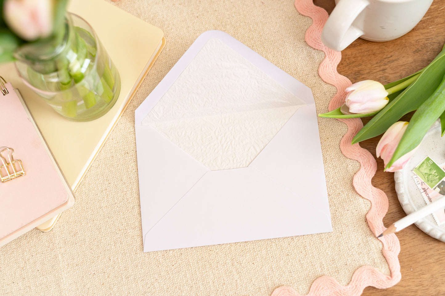 Mockup example of the Luxury Lined White Envelope that will accompany the card.  Envelope is on a table with a pink border table cloth, some tulips, a drink and stationery