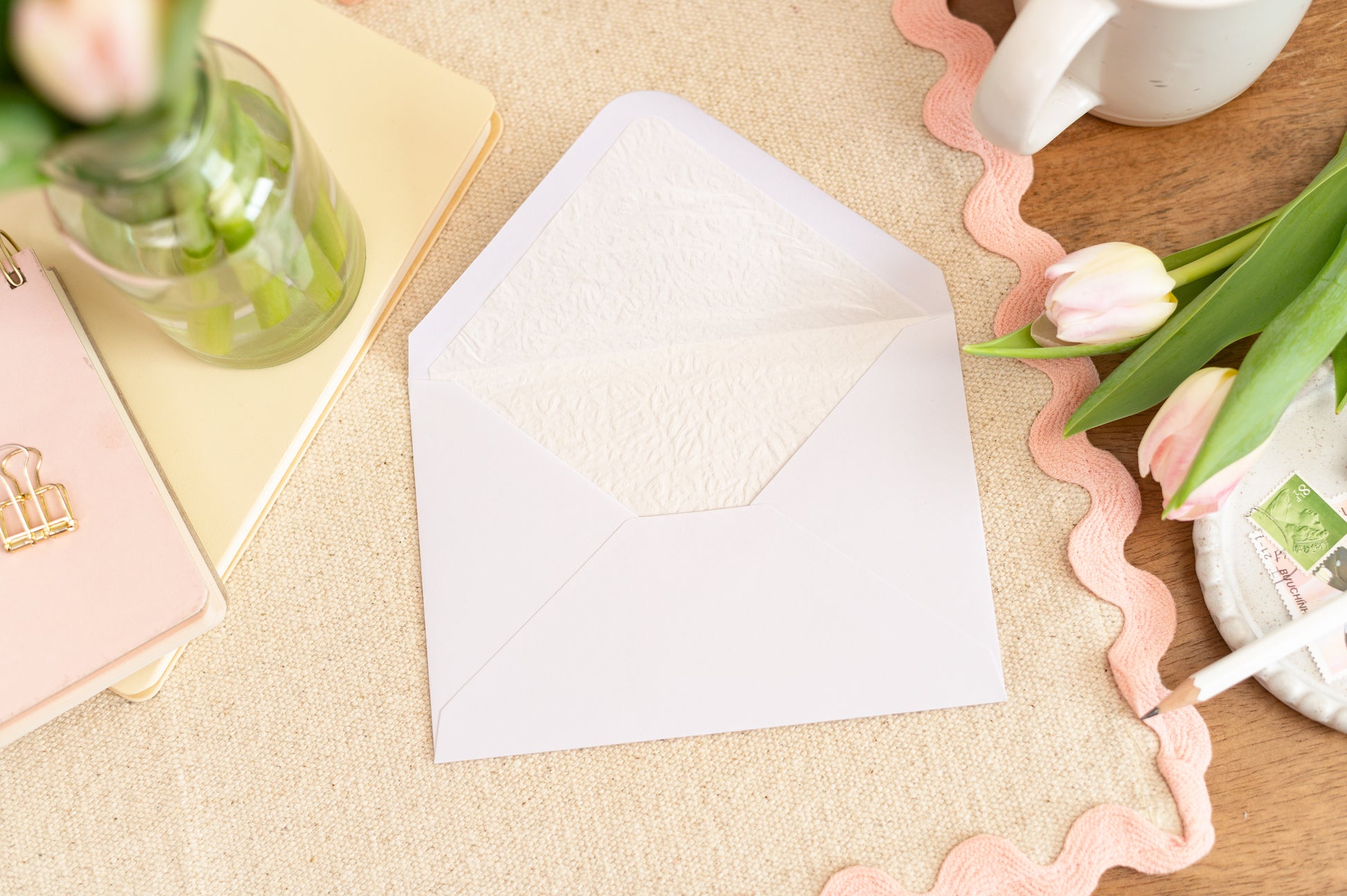 Mockup example of the Luxury Lined White Envelope that will accompany the card.  Envelope is on a table with a pink border table cloth, some tulips, a drink and stationery