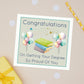Masters Degree Well Done Greeting Card. The card has an illustration of with books of different colours in the middle and mixed balloon bunches on both sides. The card has the text "congratulations on getting your degree - so proud of you"