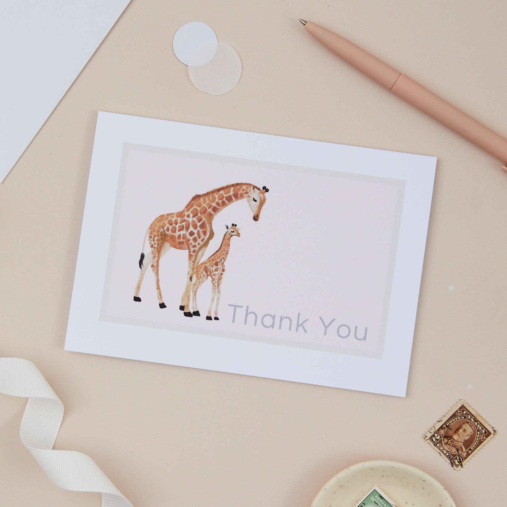 A thank you greeting card with a giraffe and its baby design, accompanied by a pen, ribbon, and decorative items on a beige background.