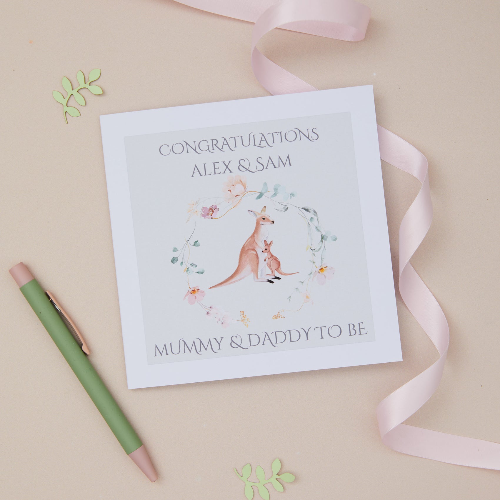 A personalized greeting card with a kangaroo and baby design, featuring the text 'CONGRATULATIONS ALEX & SAM MUMMY & DADDY TO BE'. The card is displayed alongside a white envelope and a pink ribbon on a neutral background.