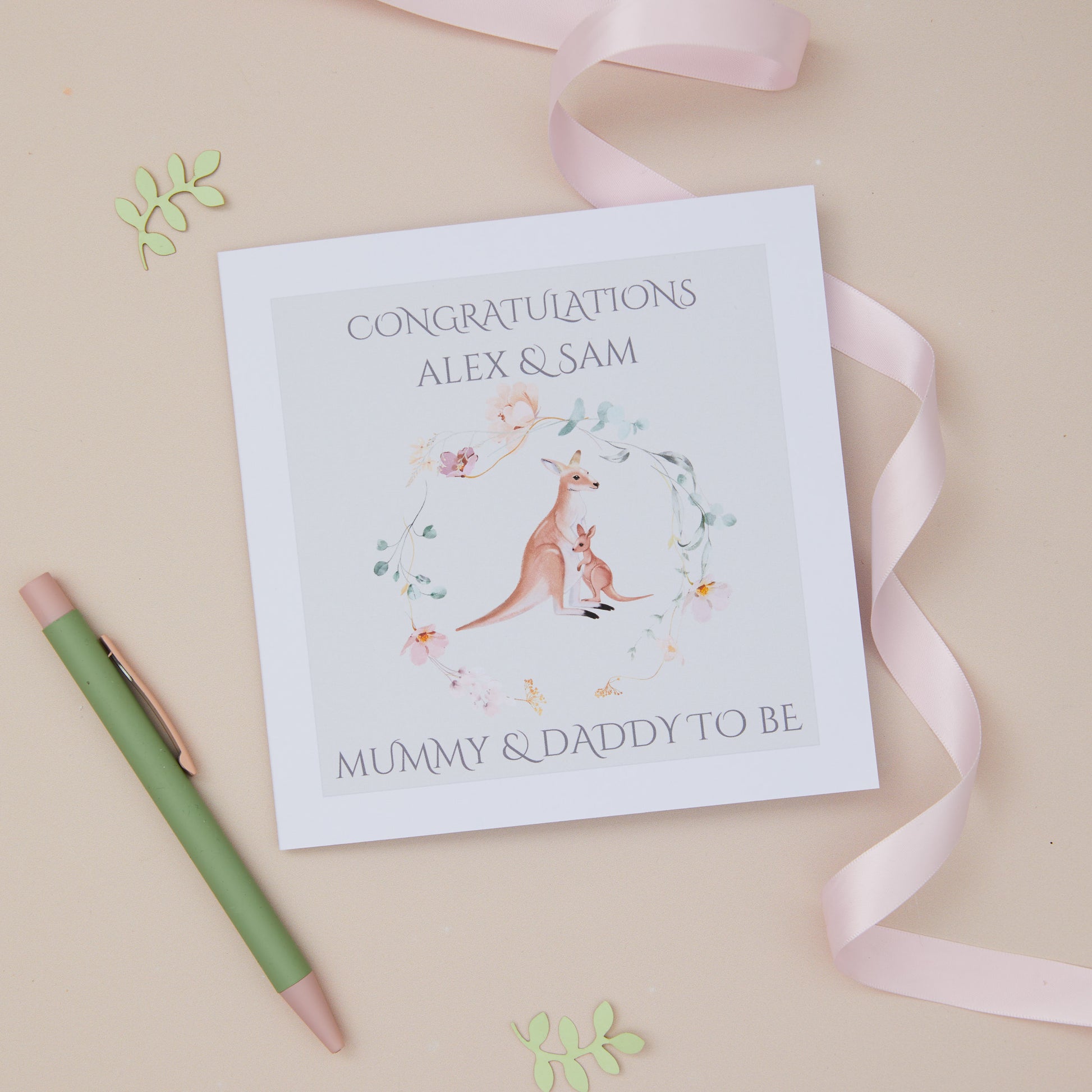 A personalized greeting card with a kangaroo and baby design, featuring the text 'CONGRATULATIONS ALEX & SAM MUMMY & DADDY TO BE'. The card is displayed alongside a white envelope and a pink ribbon on a neutral background.