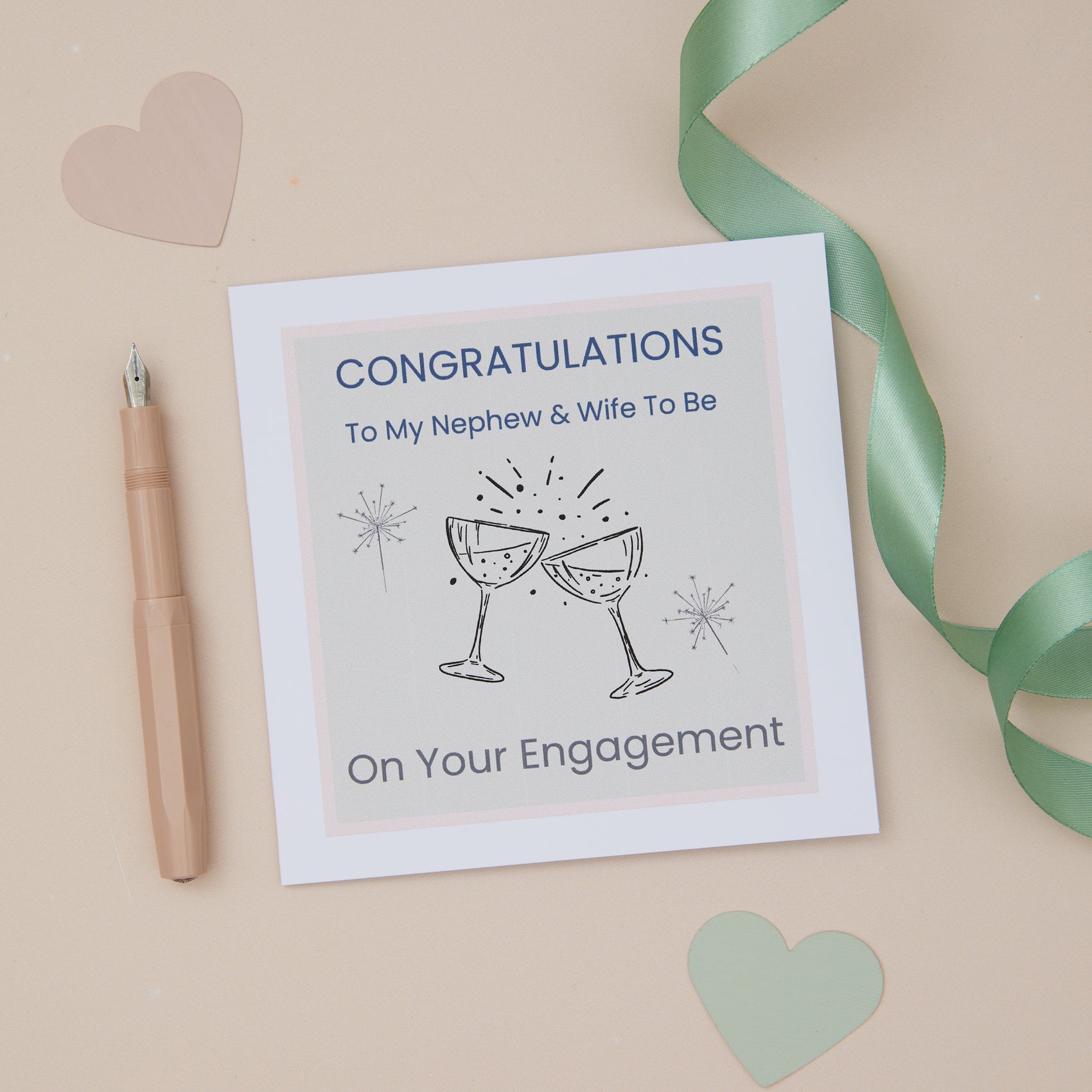 A Nephew and wife to be engagement congratulations card with two celebratory glasses design, placed on a neutral background with a pen, ribbon, and cut-out hearts.