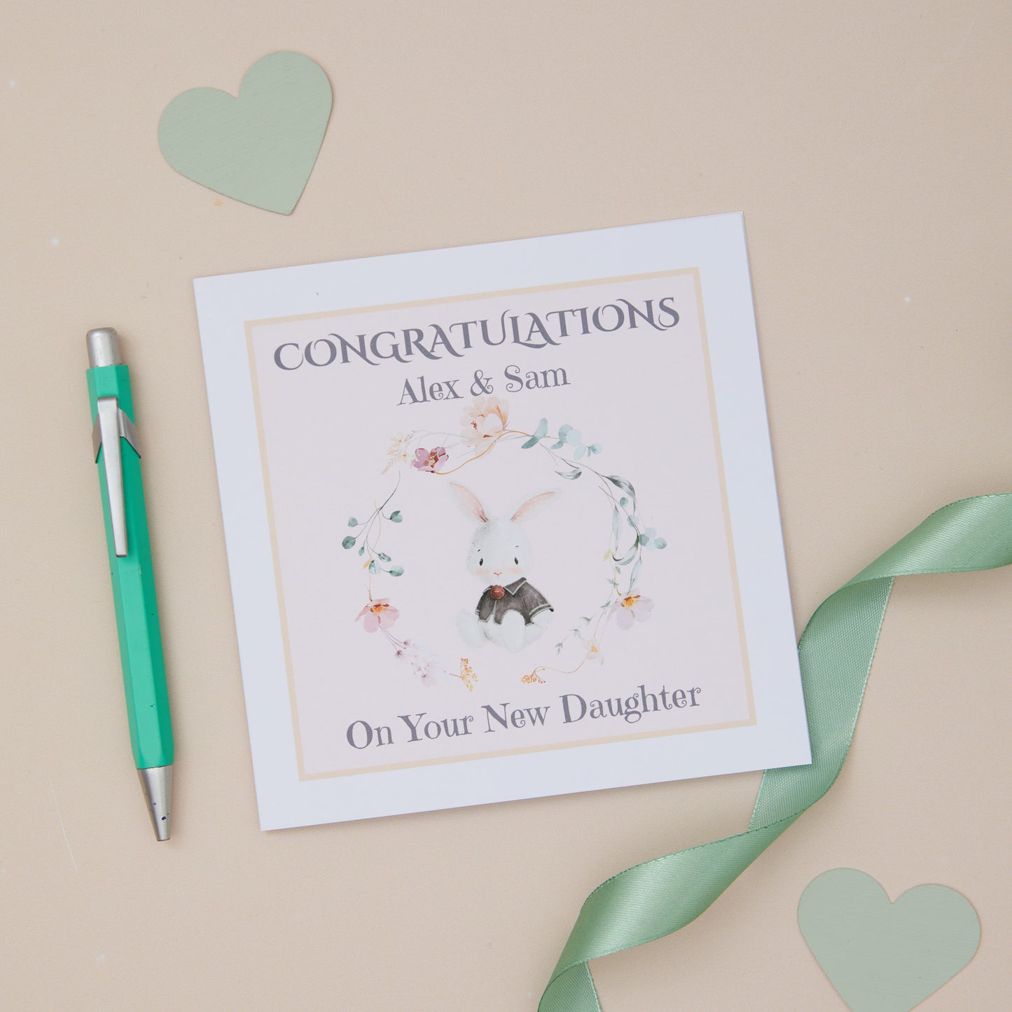 A personalised congratulations card with 'Congratulations on Your New Daughter' text, an adorable bunny illustration, and the names 'Alex & Sam'. The card is placed on a neutral background with a white envelope beside it and decorative elements like a pen and ribbon.