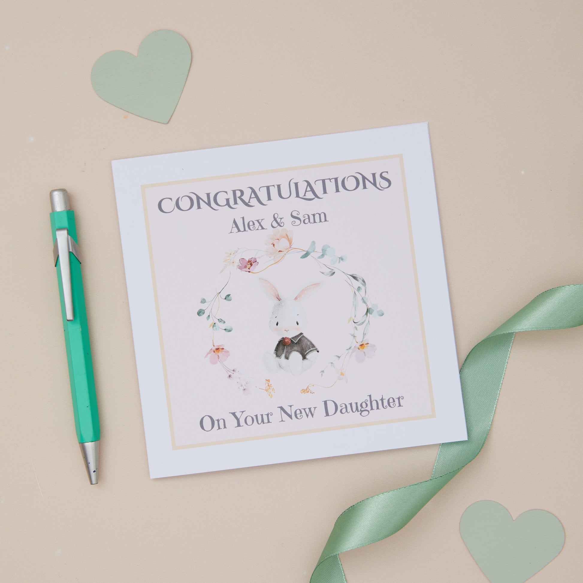 A personalised congratulations card with 'Congratulations on Your New Daughter' text, an adorable bunny illustration, and the names 'Alex & Sam'. The card is placed on a neutral background with a white envelope beside it and decorative elements like a pen and ribbon.