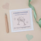 A Niece colourful engagement congratulations card with two glasses toasting, placed on a neutral background with a pen and decorative hearts.