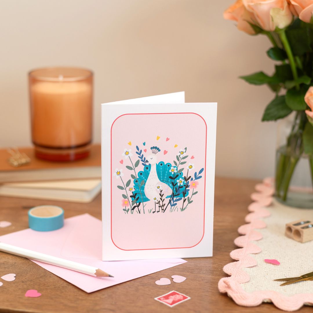 Peacock Blank Greeting Card Set with an elegant peacock design on a soft pink background, displayed on a table with a pink envelope, decorative hearts, and a floral arrangement in the background.