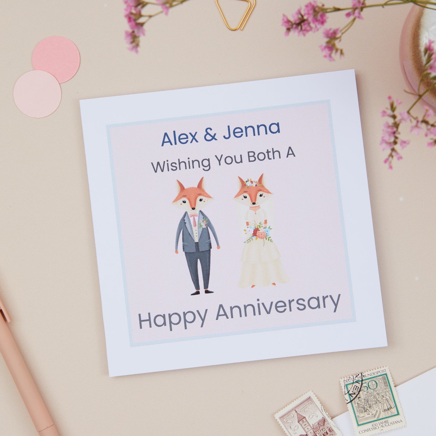 A Personalised Wedding Anniversary Card with a pink background, featuring an illustration of two foxes, a man in a suit, and a woman in a dress. The text on the card reads 'Wishing you both a Happy Anniversary' with the names 'Alex & Jenna'.