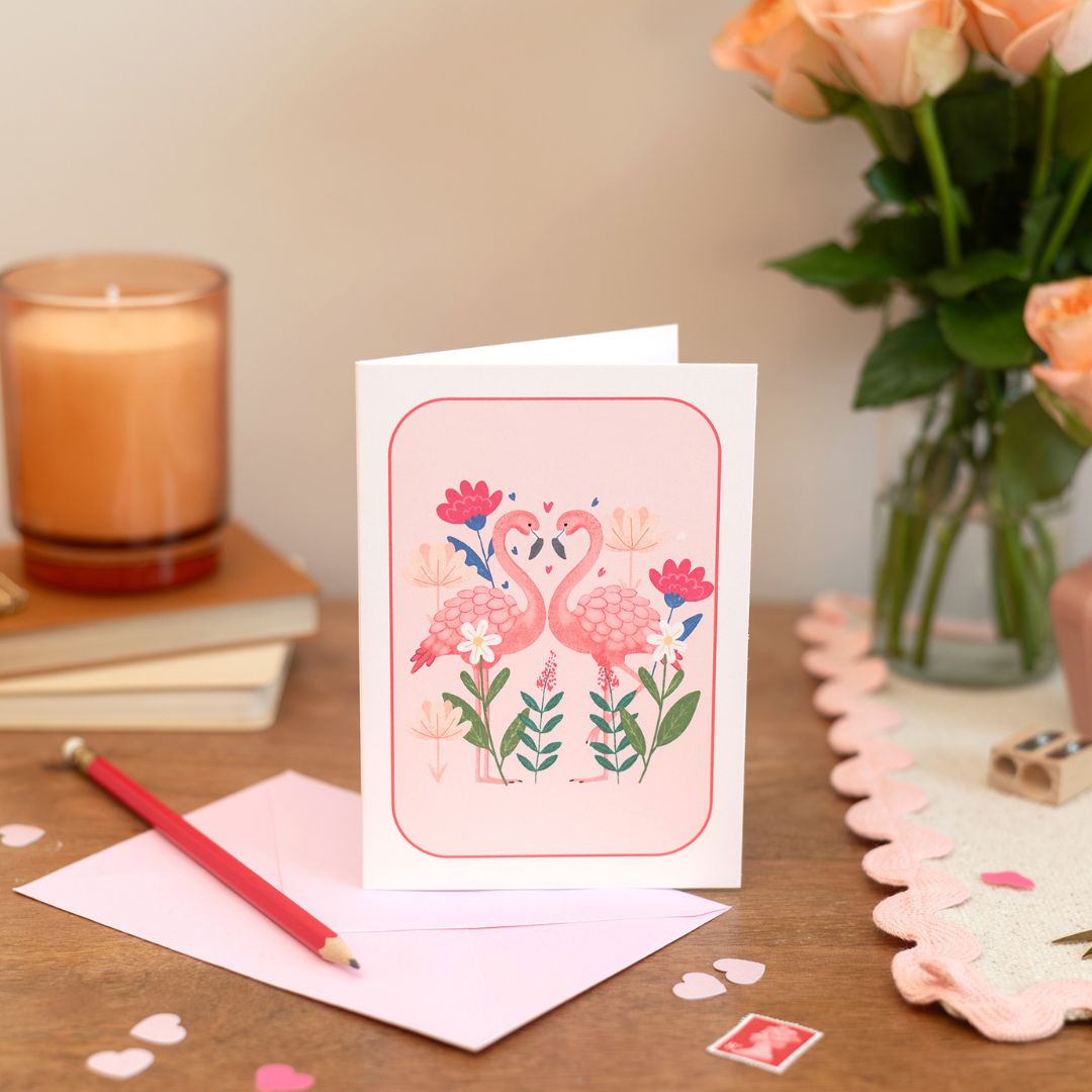 Pinks Flamingos Card Set of 8 with pink  envelopes, placed on a table with a candle and floral arrangement in the background.