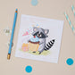 A children's Birthdaygreeting card featuring an illustrated raccoon holding a cake with a birthday present and party poopers next to it. The Card is  positioned on a table with pencils, and decorative elements.