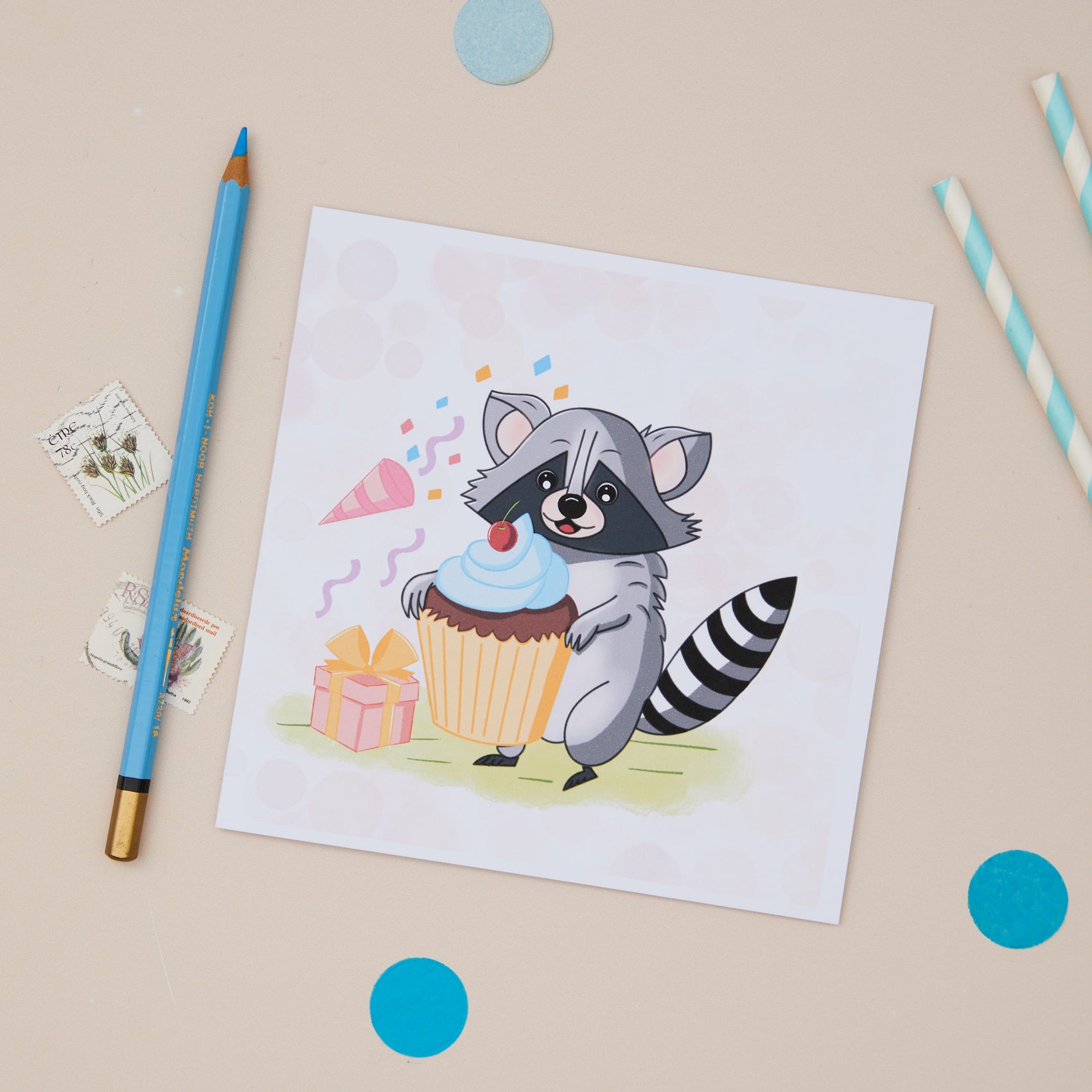 A children's Birthdaygreeting card featuring an illustrated raccoon holding a cake with a birthday present and party poopers next to it. The Card is  positioned on a table with pencils, and decorative elements.