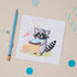 Kids Racoon Occasions greeting card featuring an illustrated raccoon holding a cake with a birthday present and party poopers next to it. The Card is  positioned on a table with pencils, and decorative elements.