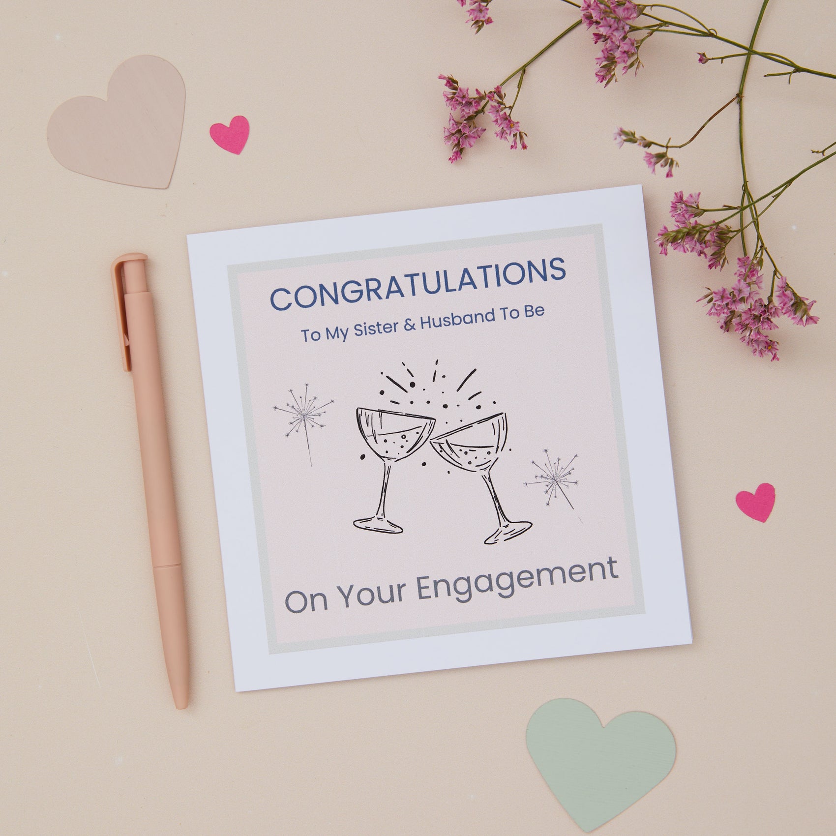 A congratulations card for a sister's engagement featuring two celebratory glasses, with a pink pen, and decorative flowers on a pastel background.