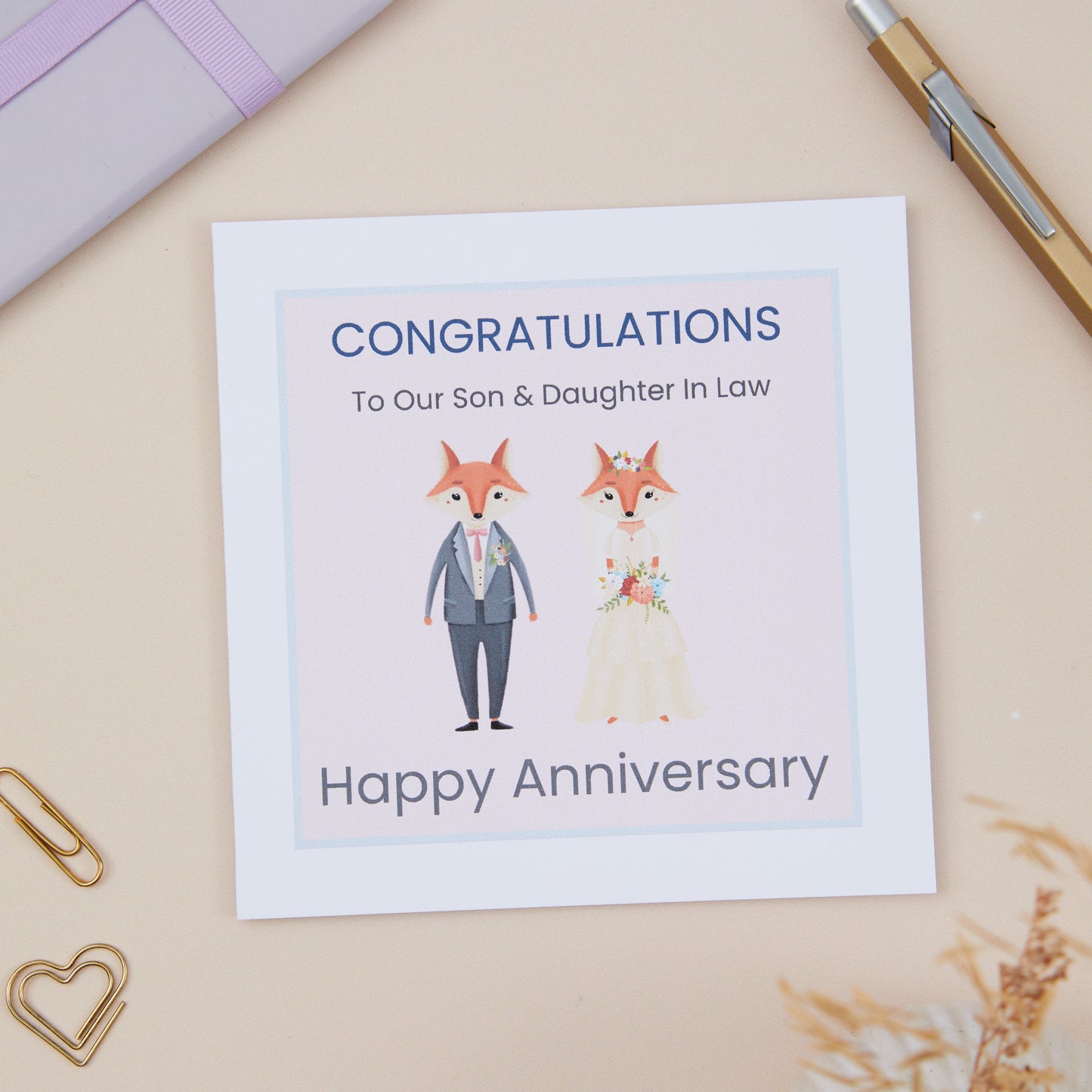 Son and Daughter In Law Anniversary Card with an illustration of a fox couple dressed in bridal attire, with the text 'CONGRATULATIONS To Our Son & Daughter in Law Happy Anniversary' on it, placed on a desk with a pen and paper clips around.