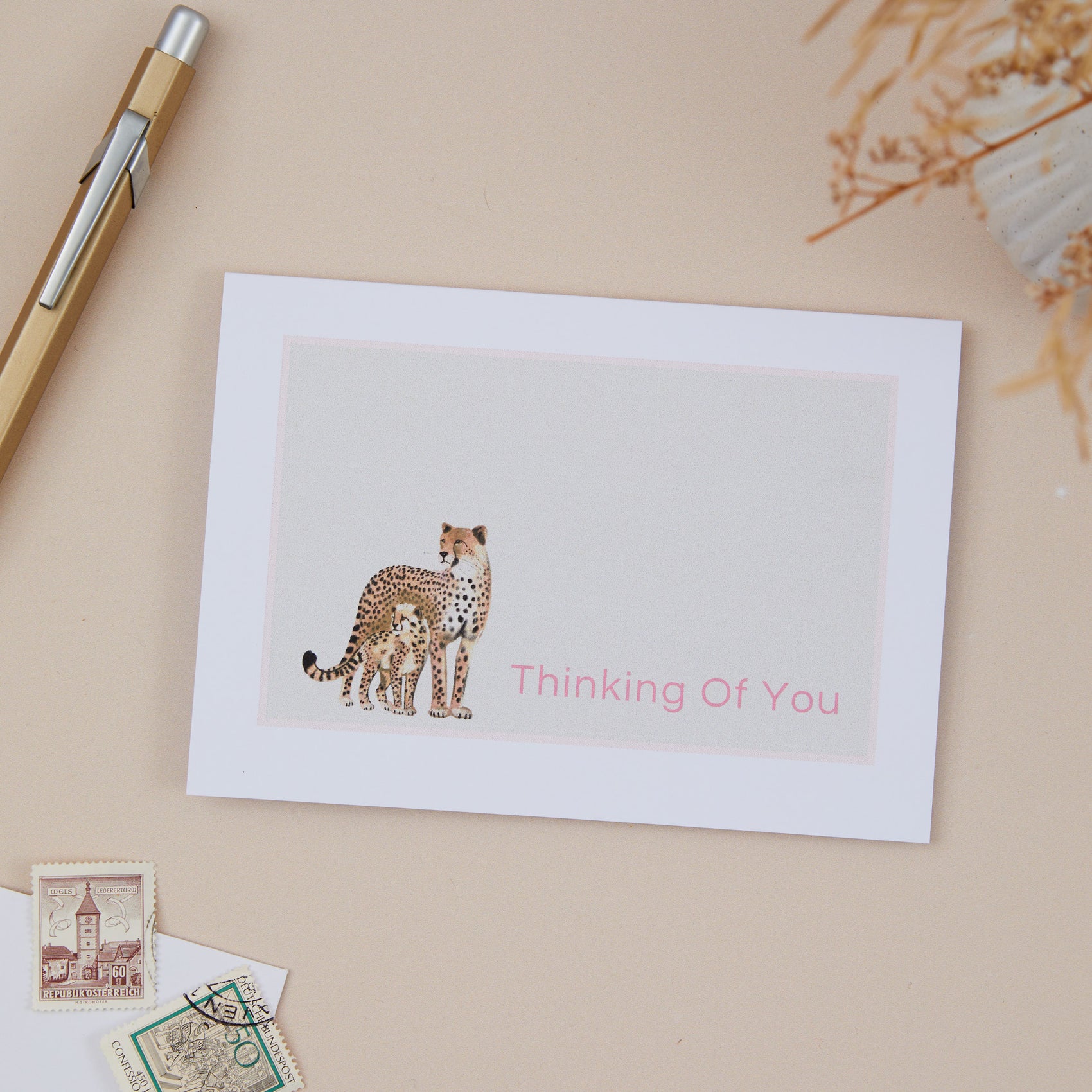 A Sympathy card with an illustration of a cheetah and its cub, with the text 'Thinking Of You' on the front. The card is placed on a beige background with a pen and some decorative elements.
