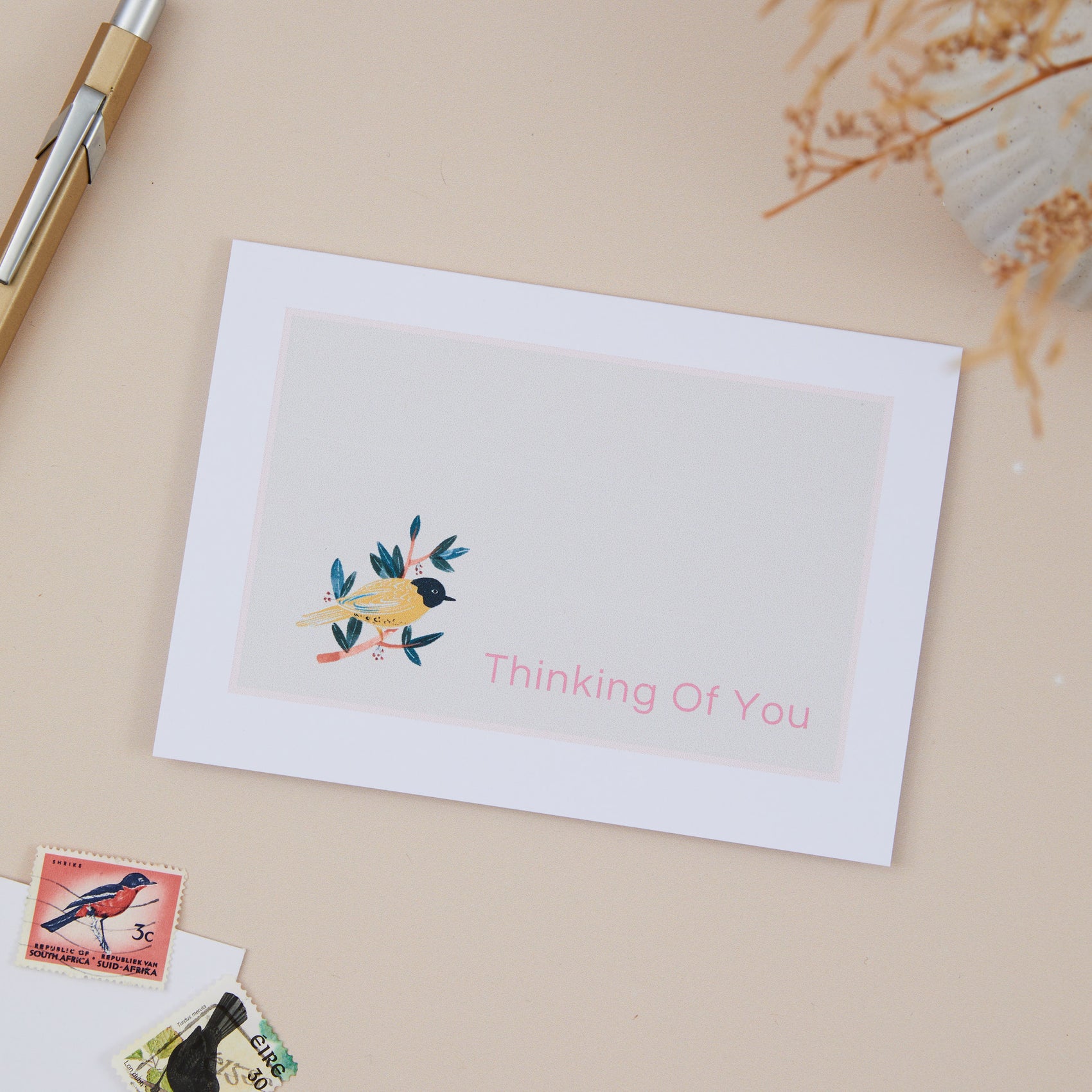 A greeting card with a watercolor illustration of a yellow bird on a branch on it. It has the text 'Thinking Of You' at the bottom, placed on a desk with a pen and some dried plants in the background.