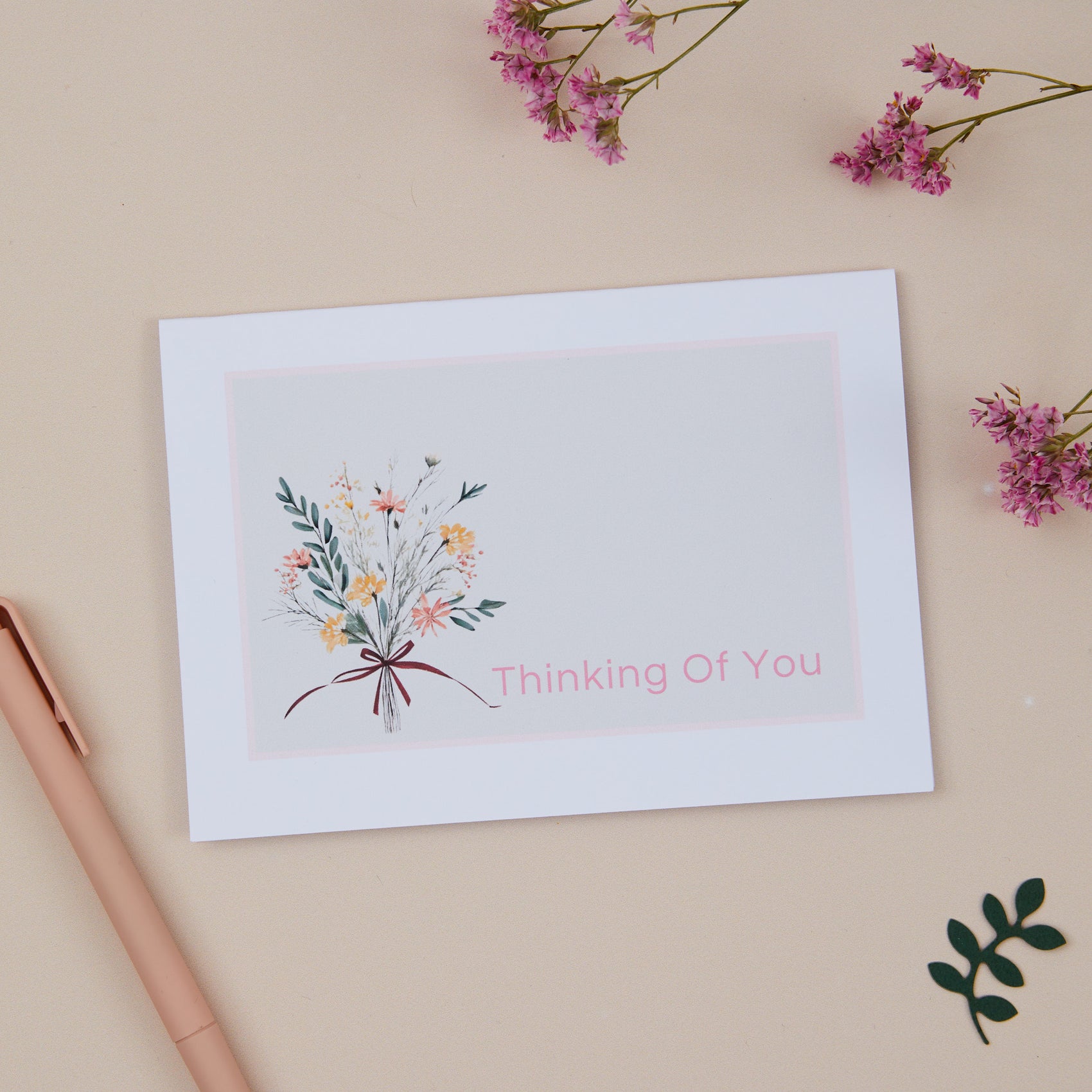 A floral sympathy card with a pastel-colored floral bouquet and the text 'Thinking Of You' on the front, placed on a neutral background with a few decorative flowers and a kraft envelope to the side.