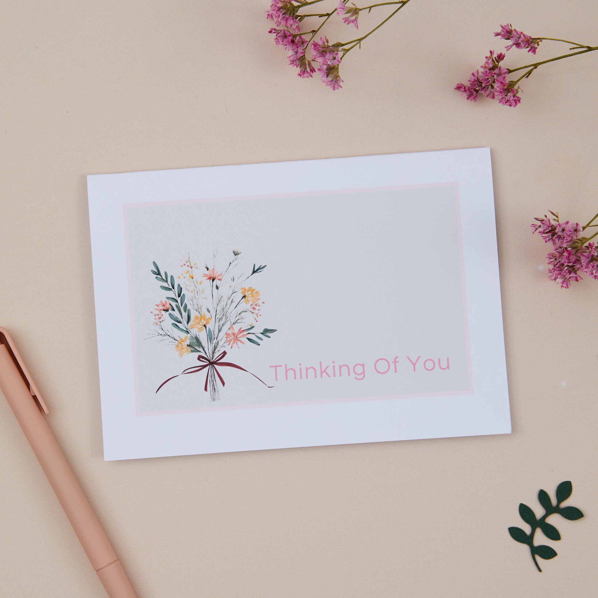 A floral sympathy card with a pastel-colored floral bouquet and the text 'Thinking Of You' on the front, placed on a neutral background with a few decorative flowers and a kraft envelope to the side.