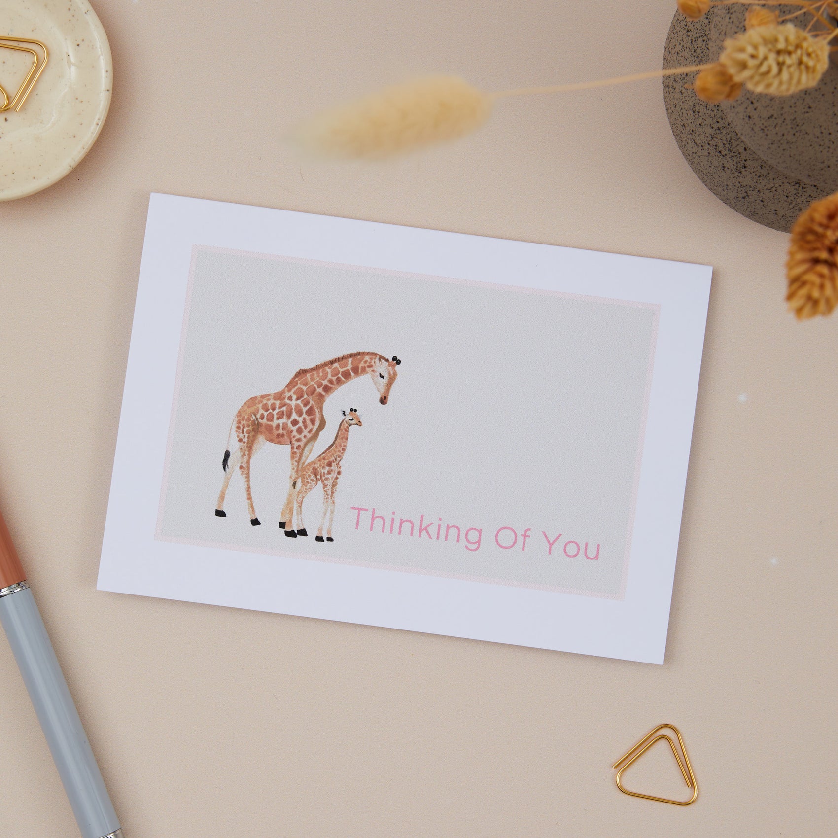 A greeting card with an illustration of a giraffe and its calf, with the text 'Thinking Of You' on the front. The card is displayed on a neutral background with decorative items, and a pen.