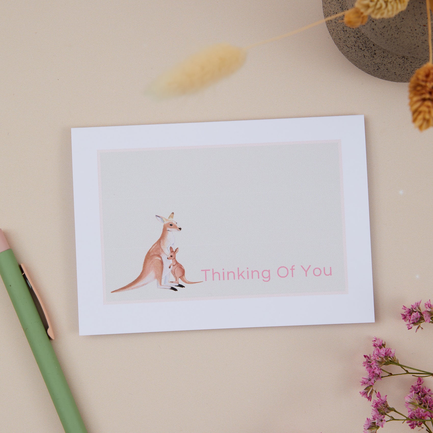 Sympathy Thinking Of You card featuring a Kangaroo and it's young. At the bottom of the card it has the words "thinking of you". The card sits on beige background with a green pen, dried flowers and other decorative eleements