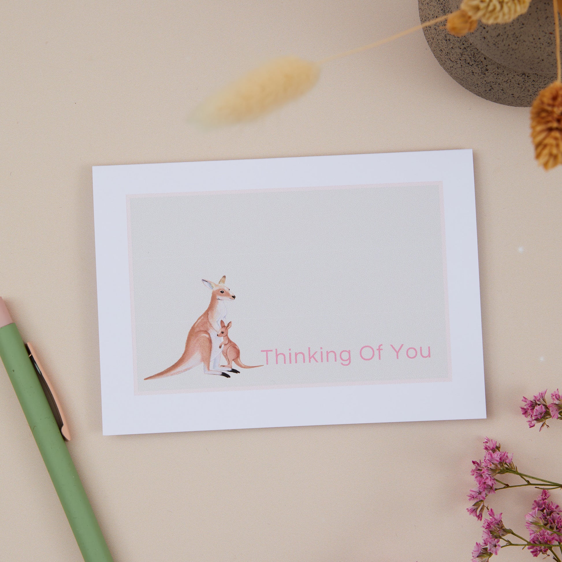 Sympathy Thinking Of You card featuring a Kangaroo and it's young. At the bottom of the card it has the words "thinking of you". The card sits on beige background with a green pen, dried flowers and other decorative eleements