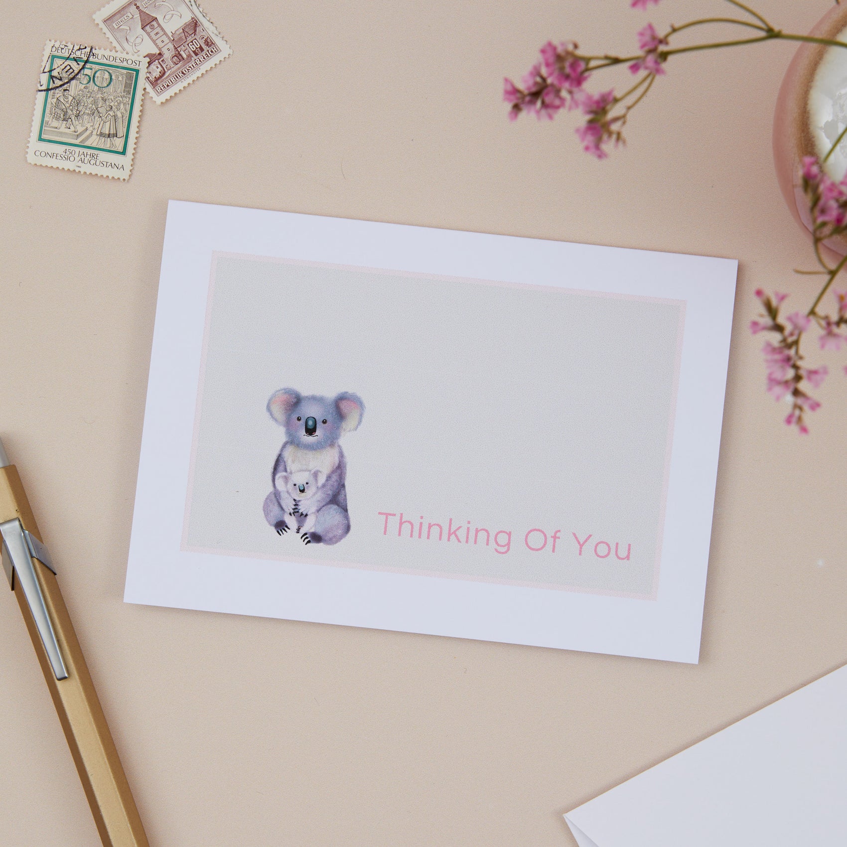 A greeting card with an image of a koala and its young, with the text 'Thinking Of You' on the card, displayed on a table with a white envelope, pens, and decorative flowers.