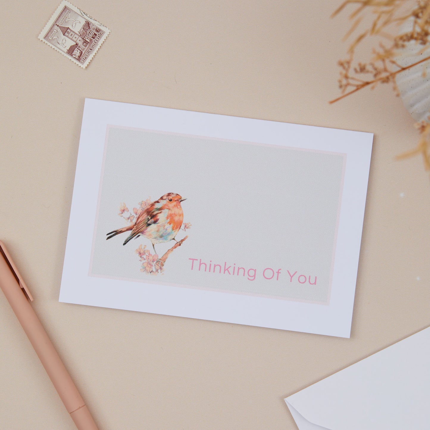 A greeting card with a red robin illustration and the text 'Thinking Of You' on the front, placed on a neutral background with a white envelope and a pen beside it.