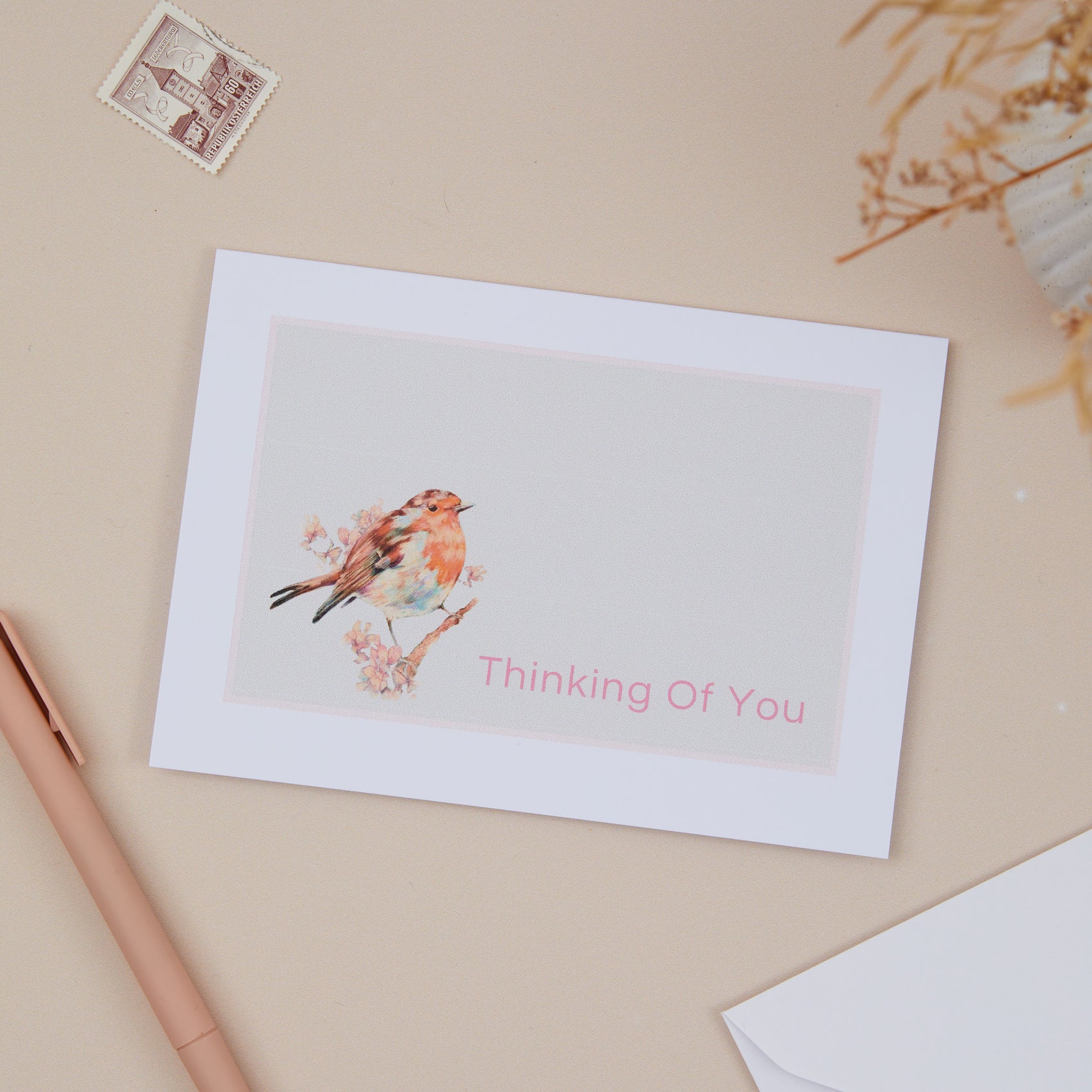 A greeting card with a red robin illustration and the text 'Thinking Of You' on the front, placed on a neutral background with a white envelope and a pen beside it.
