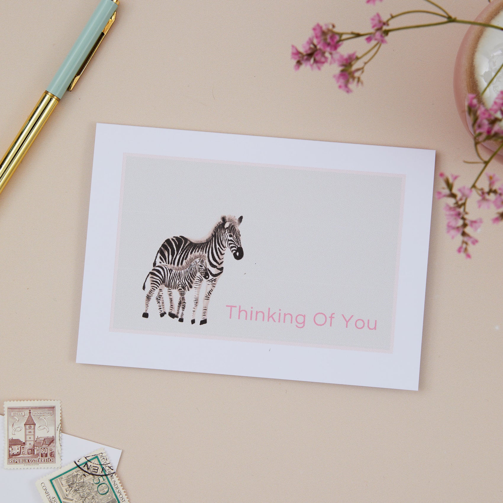 A sympathy thinking of you greeting card with an illustration of a zebra and a young zebra, with the text 'Thinking Of You' at the bottom, placed on a desk with a pen and flowers in the background.