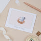 A thank you card with a watercolor illustration of a pelican sitting on an egg, the card is placed on a flat surface with a pen, ribbon, and decorative elements.