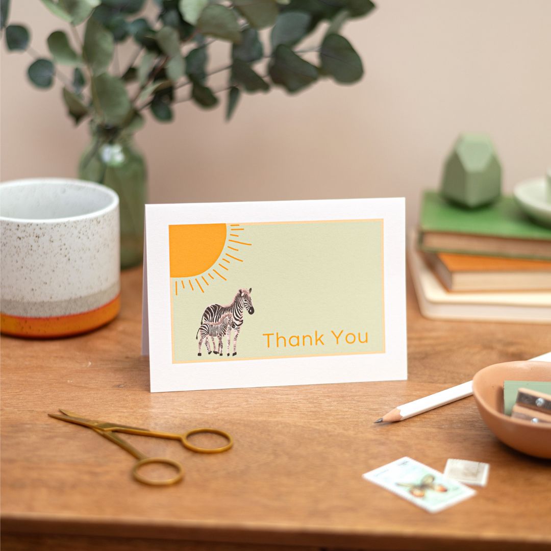A thank you greeting card with a zebra design and a sun illustration. The text on the card says "thank you". The Card is placed on a wooden table with a cup, scissors, and other stationery items in the background.