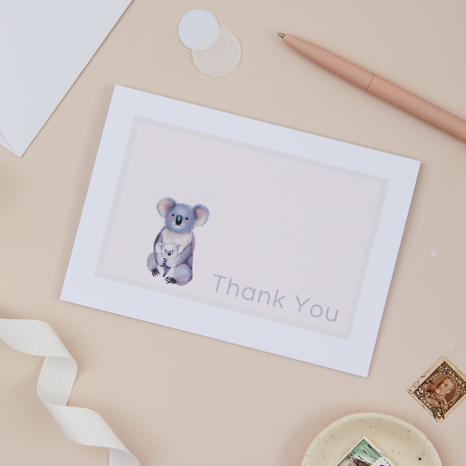 A thank you card with a koala design, placed on a desk with a pen, ribbon, and decorative elements.
