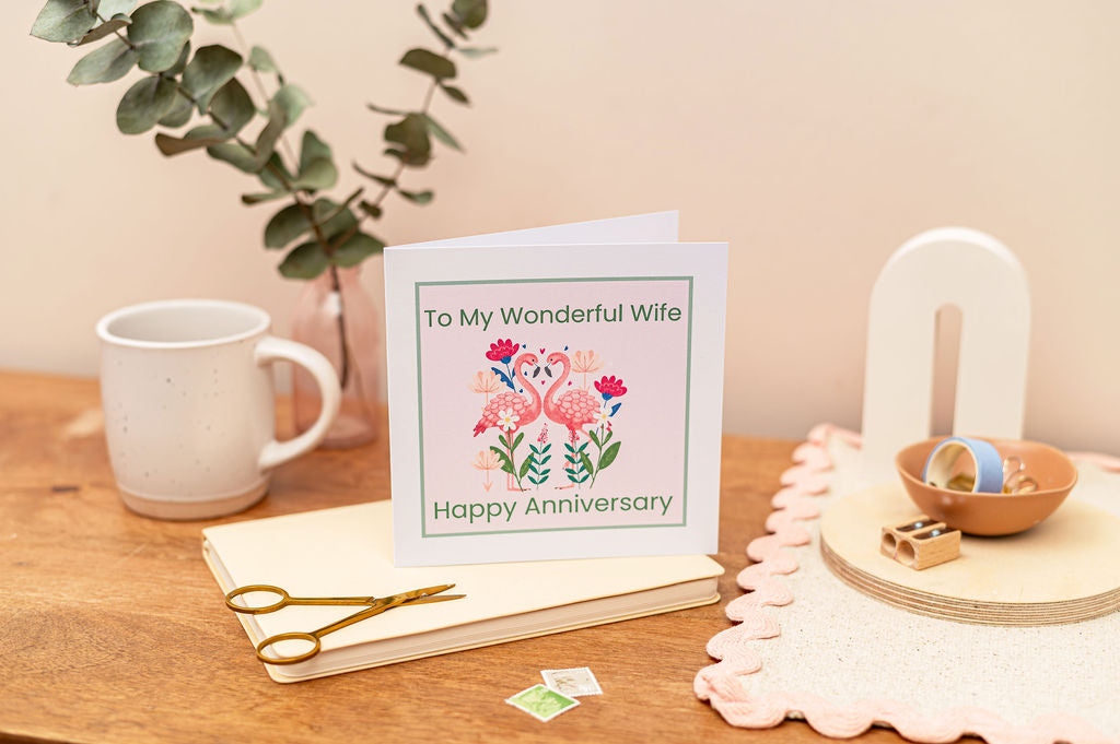 Wife Anniversary Card of two pink flamingos facing each other, the card has a soft pink background. The card is set on a desk with stationery around it.