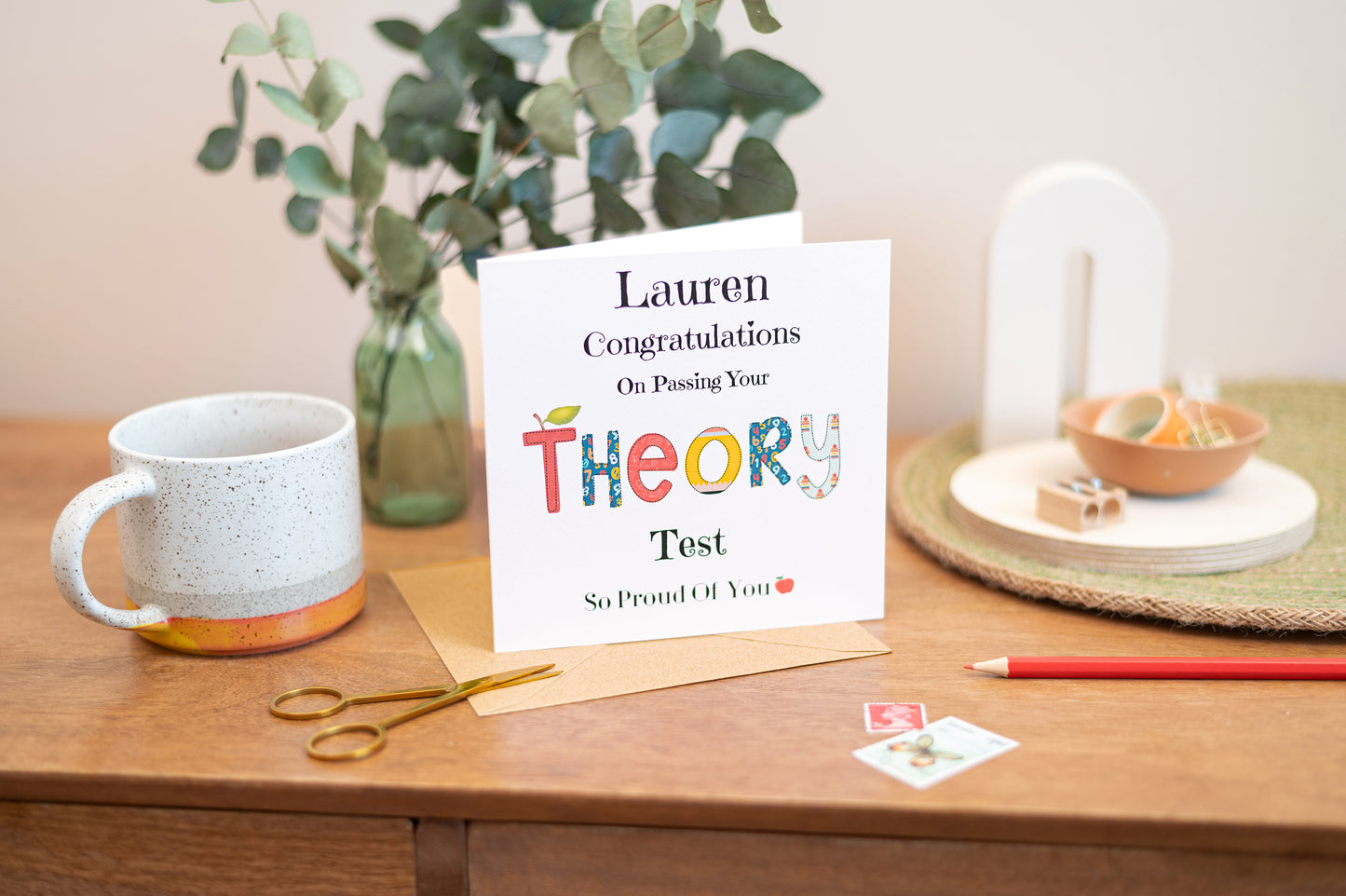 Theory Test Greeting card congratulating on passing theory test, The word theory is spelt out in a patch work type font. The card is placed on a wooden surface with a mug and stationery items.