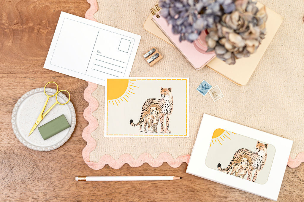 A6 Postcard with a Cheetah and it's Baby, with a yellow sun in the background. The Card is on a desk, surrounded by Stationery.