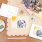 A6 Postcard with a Elephant and it's Baby, with a yellow sun in the background. The Card is on a desk, surrounded by Stationery.