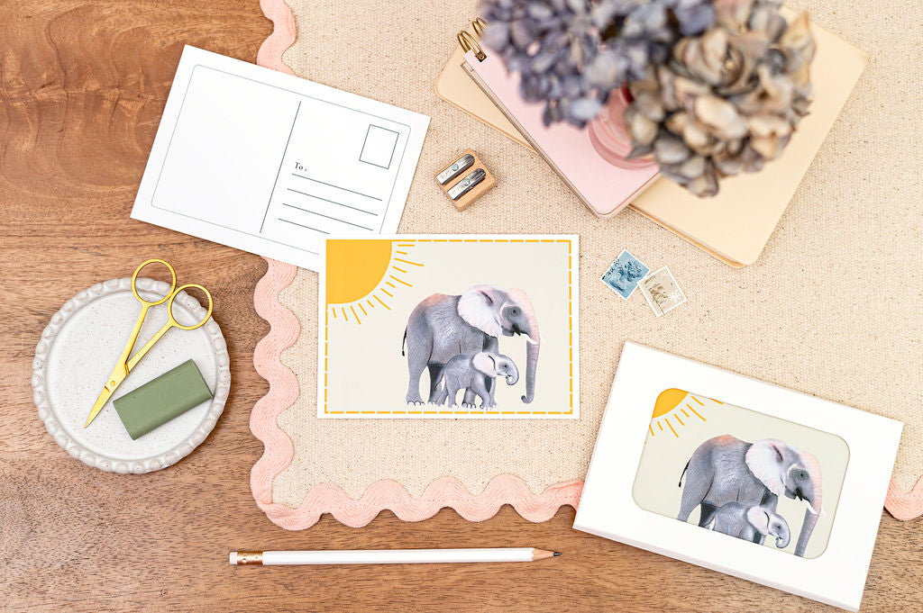 A6 Postcard with a Elephant and it's Baby, with a yellow sun in the background. The Card is on a desk, surrounded by Stationery.