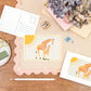 A6 Postcard with a Giraffe and it's Baby, with a yellow sun in the background. The Card is on a desk, surrounded by Stationery.