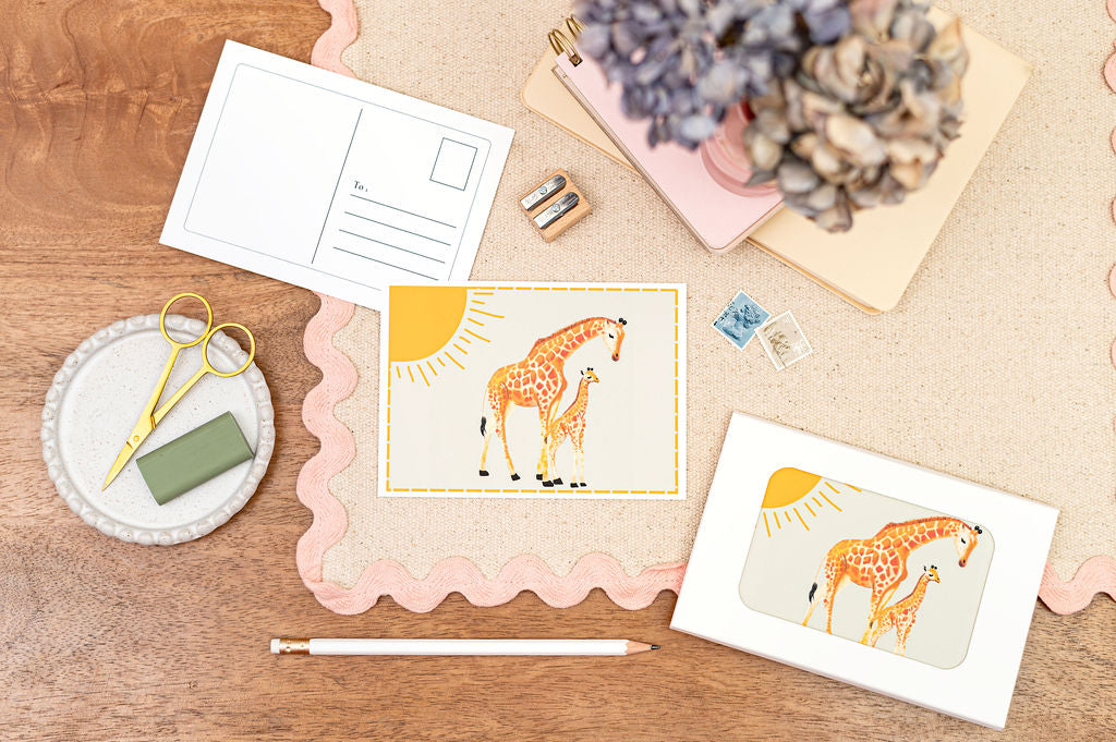 A6 Postcard with a Giraffe and it's Baby, with a yellow sun in the background. The Card is on a desk, surrounded by Stationery.