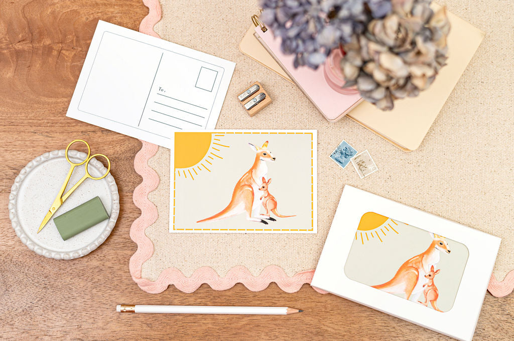 A6 Postcard with a Kangaroo and it's Baby, with a yellow sun in the background. The Card is on a desk, surrounded by Stationery.