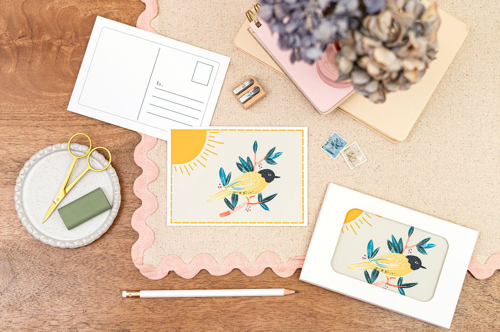 A6 Postcard Set of a Yellow Bird on a Branch. There is a yellow border on the card, and the card is on a desk with stationery.