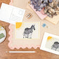 A6 Postcard with a Zebra and it's Baby, with a yellow sun in the background. The Card is on a desk, surrounded by Stationery.